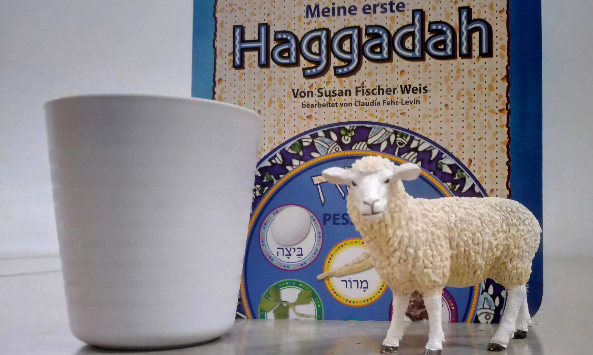 A toy sheep and a white container are standing in front of a Haggadah for children.