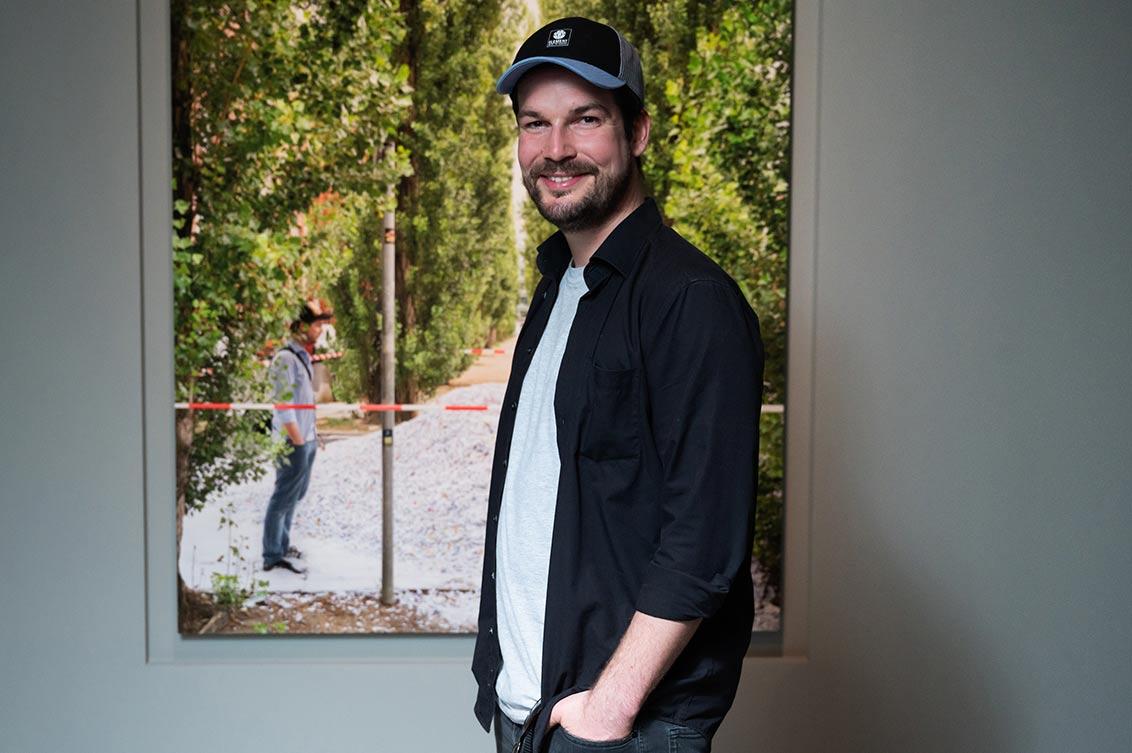 A young man with a baseball cap, beard and hands in his pockets stands in front of a portrait of himself, where he, also with a baseball cap and hands in his pockets, stands in front of a large mountain of paper scraps between green trees