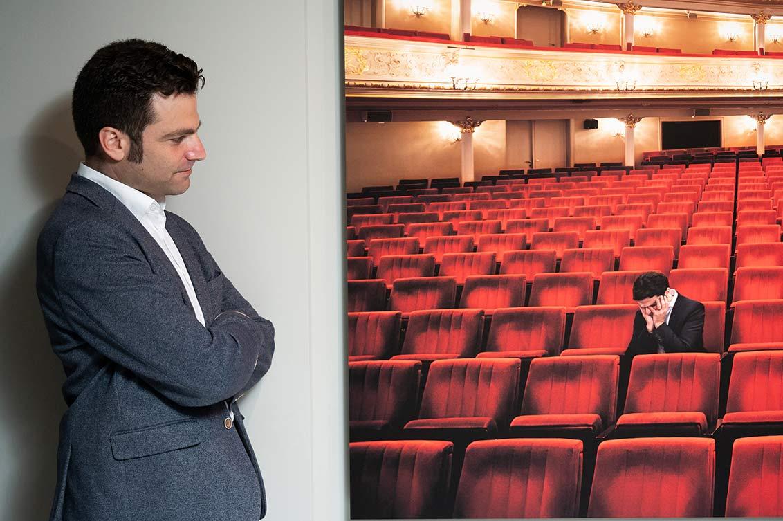 A man in a suit stands sideways next to a painting in the exhibition and looks with folded arms at the portrait of himself in the rows of seats of the Comic Opera House Berlin