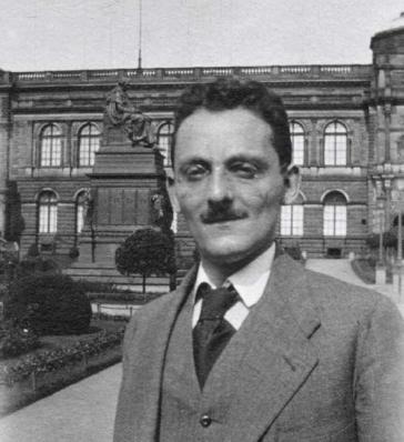 Black and white photo of a man with a moustache in a suit, a large building in the background.