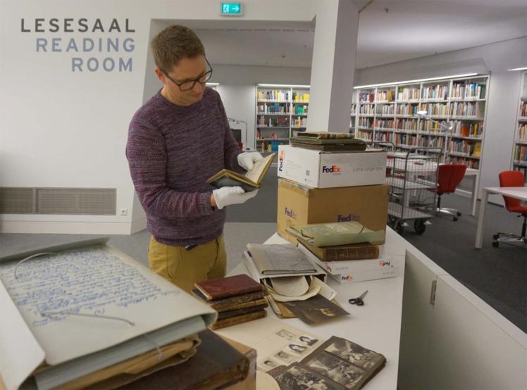 Halbausgepackte Kiste in einer Bibliothek, auf den Tisch im Vordergrund liegen Schwarz-Weiß-Fotos und weitere Dokumente, ein Archivmitarbeiter liest in einem der ausgepackten Alben