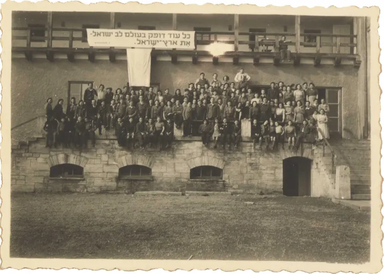 Black and white photography. About 100 women and men sit on a porch. Above them hangs a banner with a Hebrew quote from a song by Pooa Grinshpon.