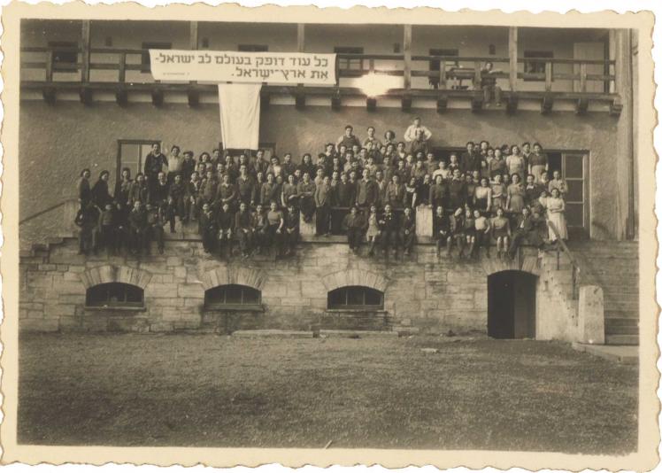 Black and white photography. About 100 women and men sit on a porch. Above them hangs a banner with a Hebrew quote from a song by Pooa Grinshpon.