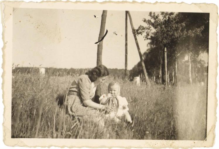 The woman wears a medium-light, patterned dress and glasses. The child sits to her right. She is wearing a light dress and a medium-light patterned headscarf (black and white photography).