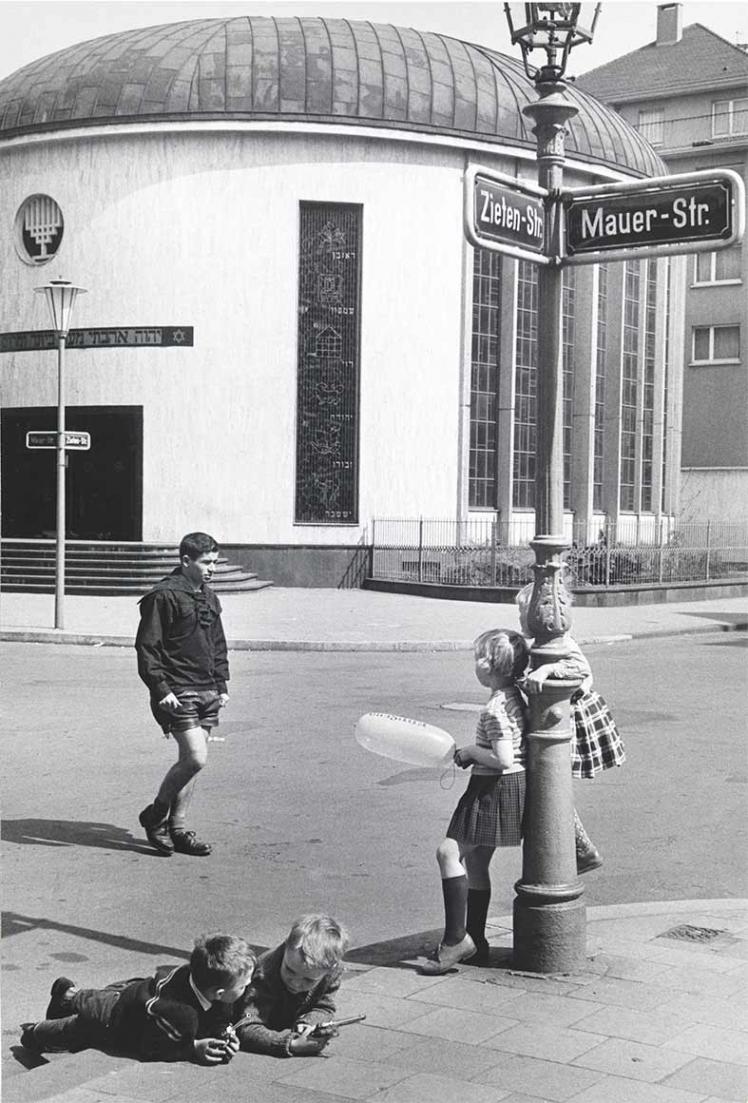 Black and white photograph: Children playing at a intersection in front of a synagogue. Two boys are lying on the curb, holding toy guns in their hands. Two girls, one of whom is holding a balloon, are standing by a street lamp. A teenager in lederhosen crosses the street.