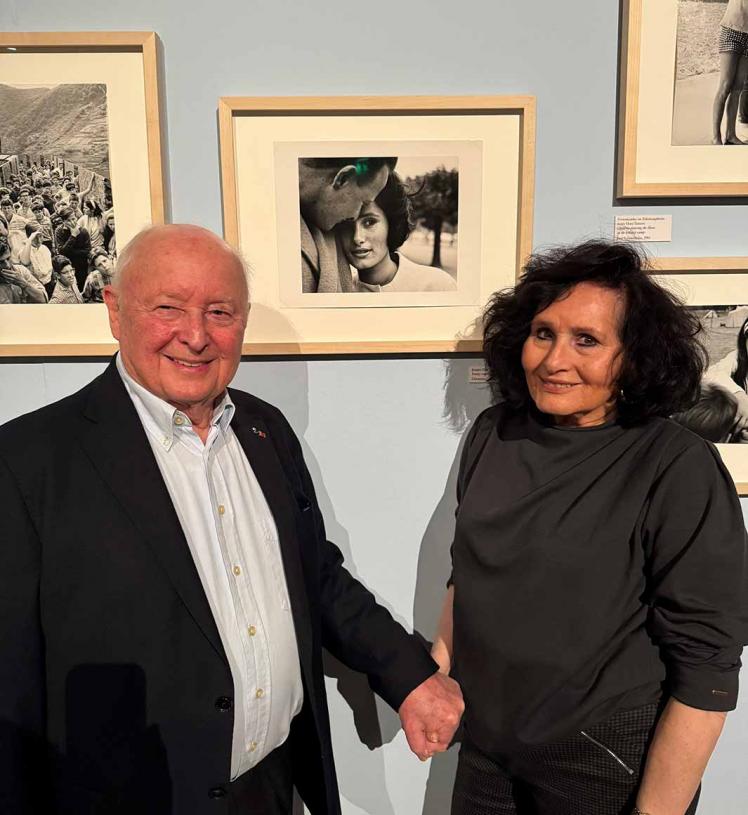 Ein älterer Mann und eine ältere Frau stehen in einer Fotoausstellung händchenhaltend vor einem Schwarz-Weiß-Foto, auf dem sie als junges Paar abgebildet sind.