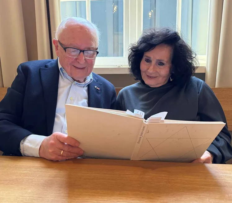 Ein Mann und eine Frau sitzen an einem Tisch und betrachten zusammen eine Seites eines aufgeschlagenen Buches, beide lächeln.