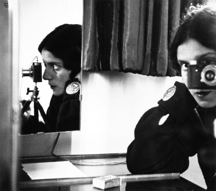 Black and white Self-Portrait of a woman with dark hair with a camera, a profile of her face can be see in the mirror sitting next to her