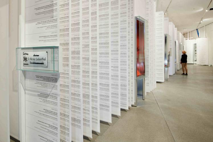 Long white paper banners with text hanging from the ceiling interrupted by showcases, in the background stands a woman with blonde hair