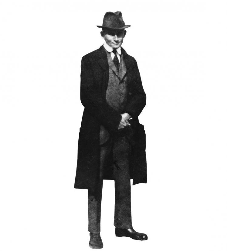 Full-length portrait of Franz Kafka, standing with coat, hat and tie, hands clasped in front of his body.