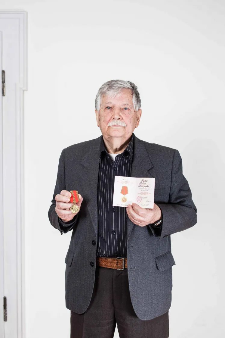 Older gentleman in dark jacket holds medal and a document in his hands