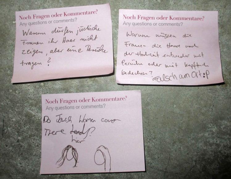 Pinke Post-it-Zettel mit Fragen und Kommentare zur Kopfbedeckung: „Warum dürfen jüdische Frauen ihr Haar nicht zeigen, aber eine Perücke tragen?“, „Warum müssen die Frauen die Haare nach der Hochzeit entweder mit Perücke oder mit Kopftuch bedecken?“