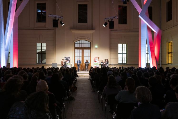  Im Glashof im Jüdischen Museum Berlin sind die Zuschauerränge gefüllt. Erhöht stehen Menschen an Pulten. Die Balken sind bunt beleuchtet.