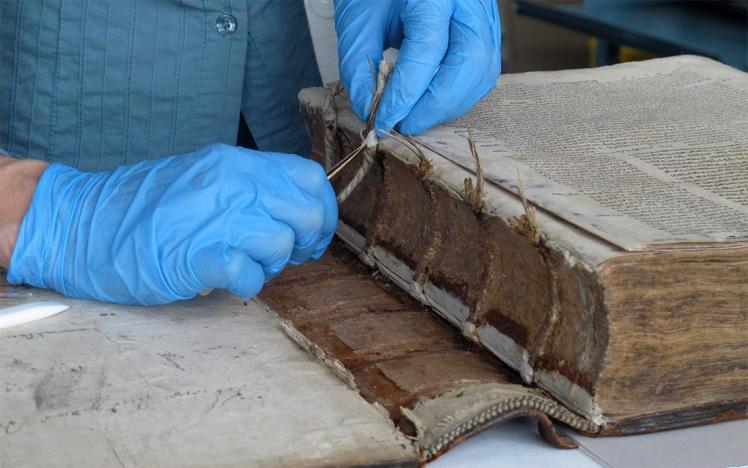 Hands in blue gloves work with a pair of tweezers on a very old, thick book