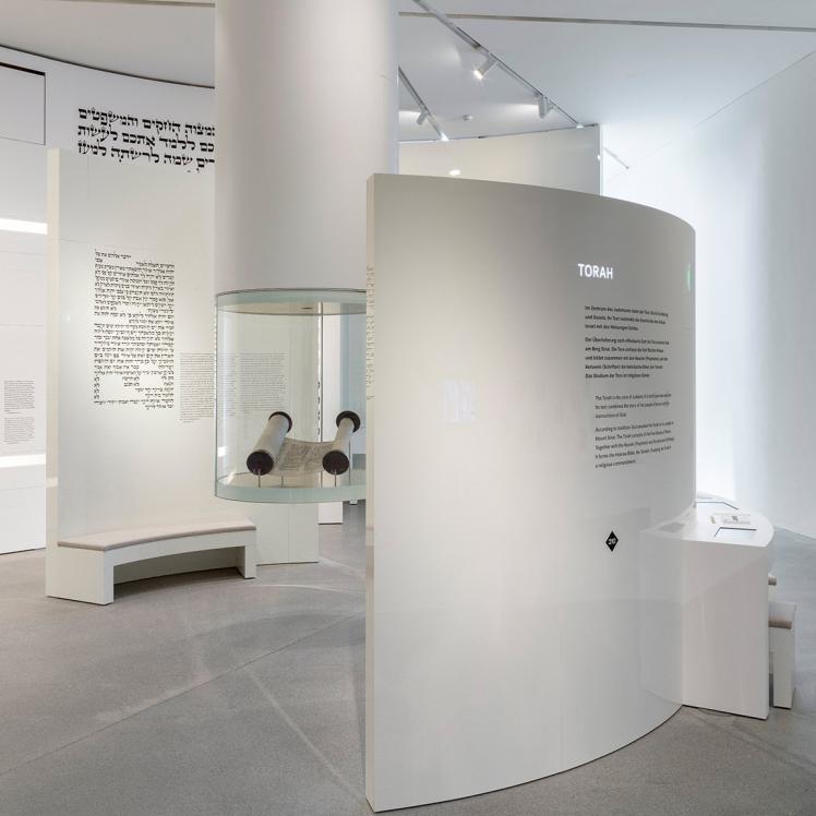 Von der Decke hängende runde Vitrine mit einer Tora-Rolle darin, an einer Wand dahinter die zehn Gebote auf Hebräisch, darum halbrunde weiße Ausstellungswände mit Text