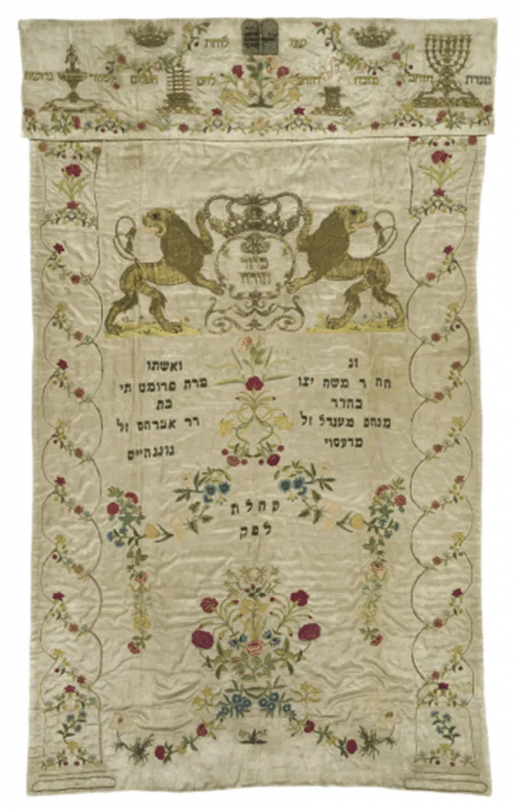 Embroidered white Torah curtain with numerous decorations and two lions and a crown over the Hebrew dedication inscription.